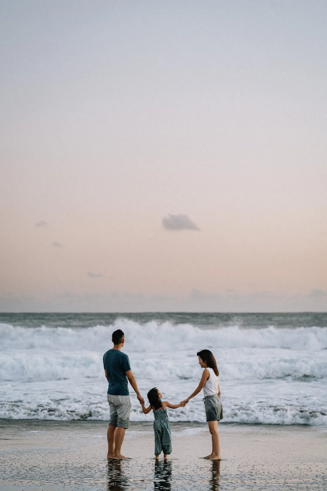 Au bord de l'eau, une petite fille tient la main de ses parents face aux vagues, sous un ciel rose