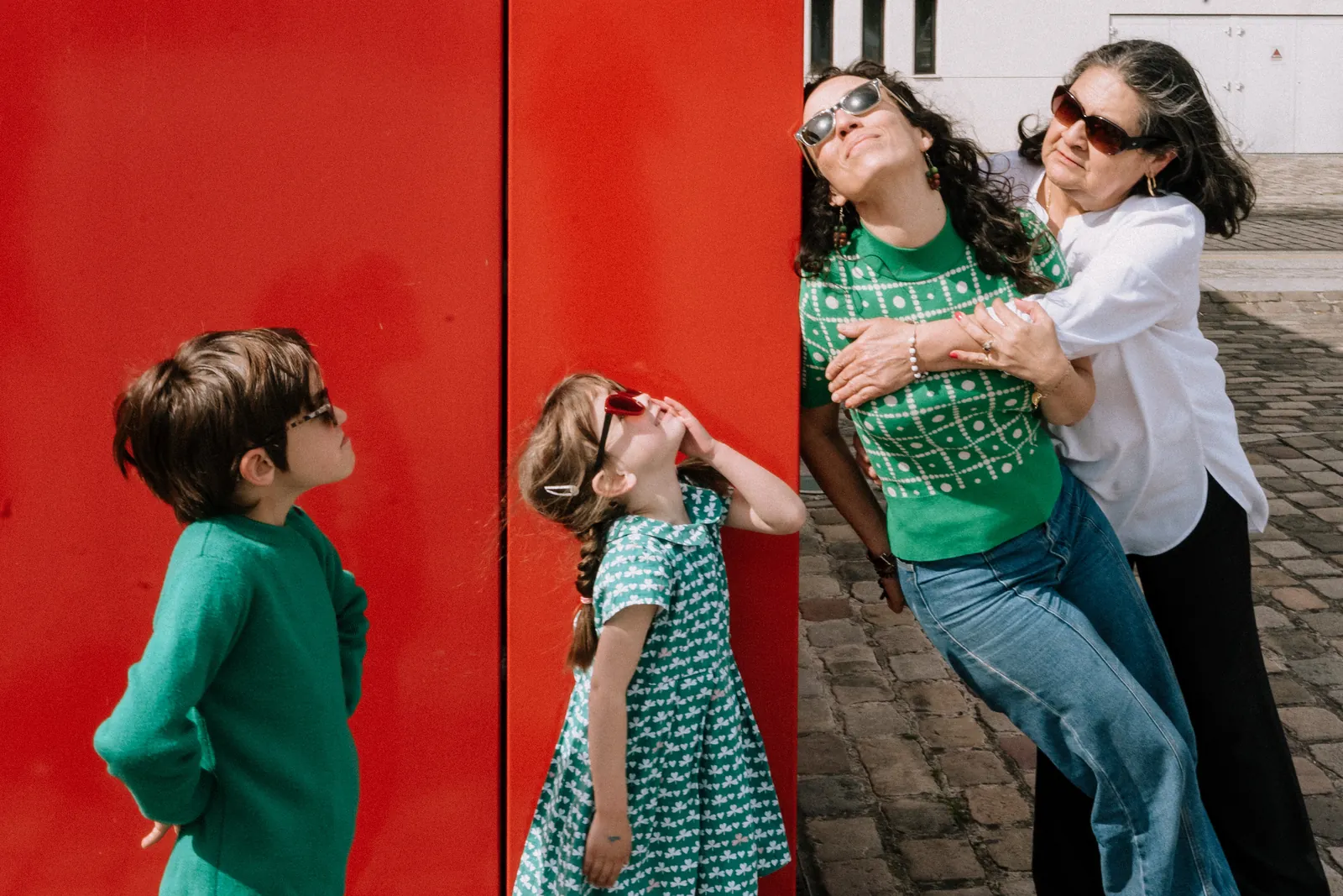 Devant un mur rouge, deux enfants à lunettes regardent le ciel pendant que maman et mamie se serrent en riant
