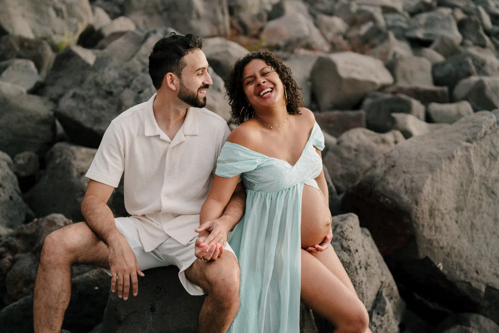 Assis sur les galets, un futur papa et une future maman en robe bleu ciel éclatent de rire, main dans la main