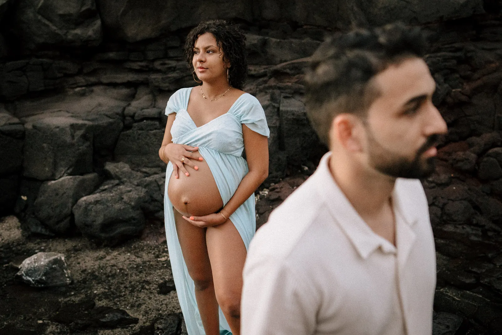 Devant les roches volcaniques, une future maman en robe bleu ciel tient son ventre, le futur papa flou au premier plan