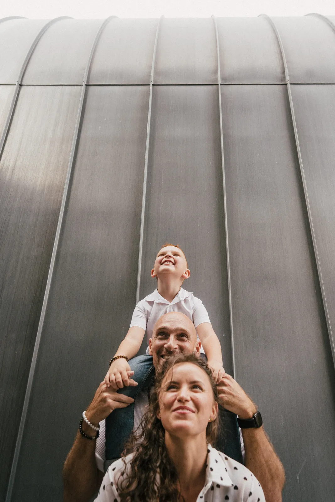 Le fils sur les épaules de son papa, la maman devant : la famille empilée en totem contre une façade de métal